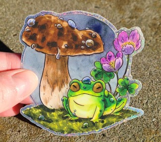 A glittery vinyl sticker of a little green frog happily sitting beneath a mushroom.  There are raindrops on the frog, mushroom, and on the sorrel flowers behind.